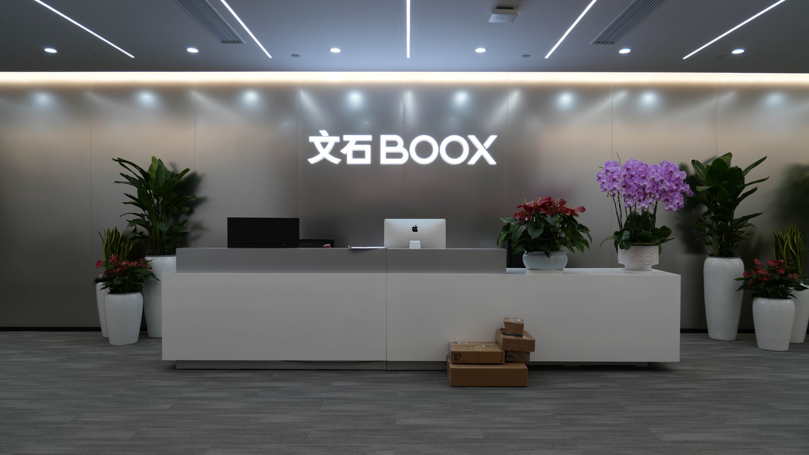 文石 BOOX 辦公室前台,灰色金屬牆面上有發光的「文石BOOX」字樣,兩側擺放盆栽與蝴蝶蘭