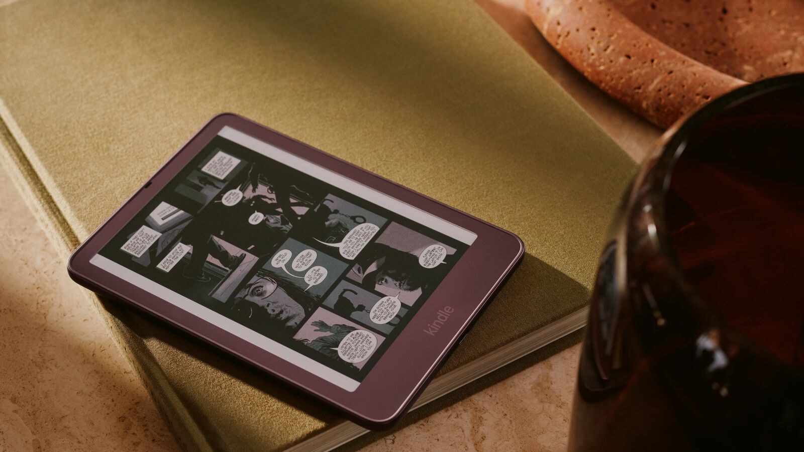 Amazon 官方情境圖:一台無花果紫紅色的 Kindle Colorsoft 放在綠色書本上,螢幕顯示彩色漫畫,旁邊有一杯飲料