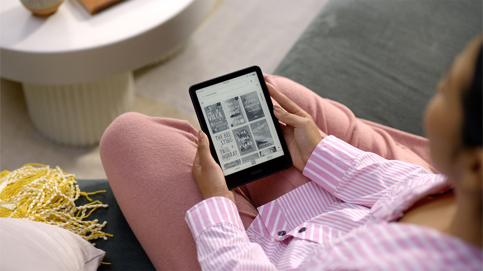 Amazon 官方情境圖:穿粉色條紋上衣的女子靠在沙發上,雙手拿著 Kindle Paperwhite 瀏覽書庫